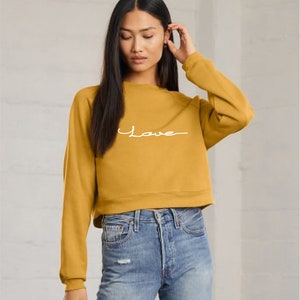 May include: A mustard yellow cropped sweatshirt with long sleeves and the word "Love" written in white cursive on the front.