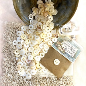 May include: A collection of vintage mother-of-pearl buttons in various sizes, spilling from a ceramic bowl onto a lace-trimmed cloth. A small paper envelope holds a button, and a vintage label reads "Mississippi River Mother of Pearl Buttons."