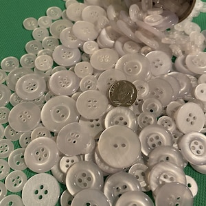 May include: A collection of various sizes of white buttons, some with four holes and others with two, spilling from a cardboard tube. A US dime is included for size reference. The buttons are scattered on a green surface.