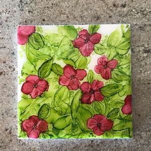 May include: A square coaster with a floral design. The coaster features pink flowers with green leaves on a white background.