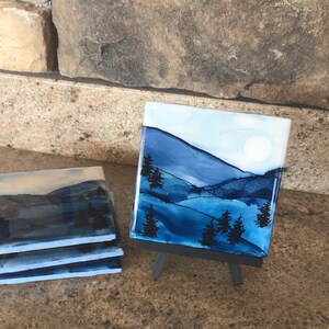 Puede incluir: Un juego de cuatro posavasos de cerámica azul y blanco con una escena de montaña de acuarela. Los posavasos están sobre una superficie marrón y uno se muestra en un pequeño caballete negro.
