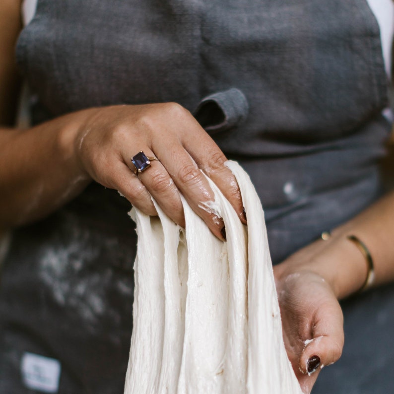 Peut inclure: Une main tenant une longue et fine bande de p&acirc;te blanche. La main porte une bague en or avec une pierre pr&eacute;cieuse violette fonc&eacute;e.