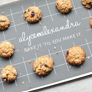 May include: A grey silicone baking mat with a white grid pattern and the text "alycealexandra bake it 'til you make it".  There are nine oatmeal cookies with cranberries on the mat.