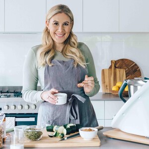 May include: A woman wearing a grey apron stands in a kitchen, holding a small white bowl and a piece of food. She is smiling at the camera. There are vegetables, a bowl of brown sugar, and a kitchen counter in the background.