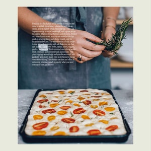 Peut inclure: Gros plan sur un pain focaccia garni de tomates cerises et de brins de romarin. La focaccia est dans un plat &agrave; p&acirc;tisserie sur une surface grise.