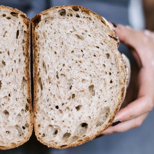 Peut inclure: Gros plan d'une miche de pain tranch&eacute;e, montrant la mie moelleuse et a&eacute;r&eacute;e et la cro&ucirc;te l&eacute;g&egrave;rement dor&eacute;e. Le pain est tenu dans une main, r&eacute;v&eacute;lant la texture et les d&eacute;tails du produit cuit.