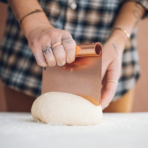 May include: A person using a copper dough scraper to cut a ball of white dough. The dough is on a white surface.