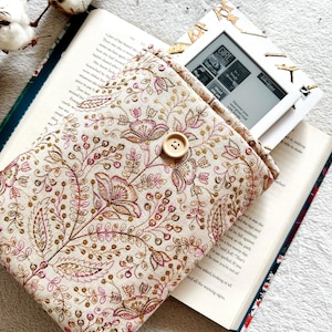 May include: A beige fabric e-reader case with a floral pattern in shades of pink and gold. The case has a button closure and is shown with an e-reader inside.