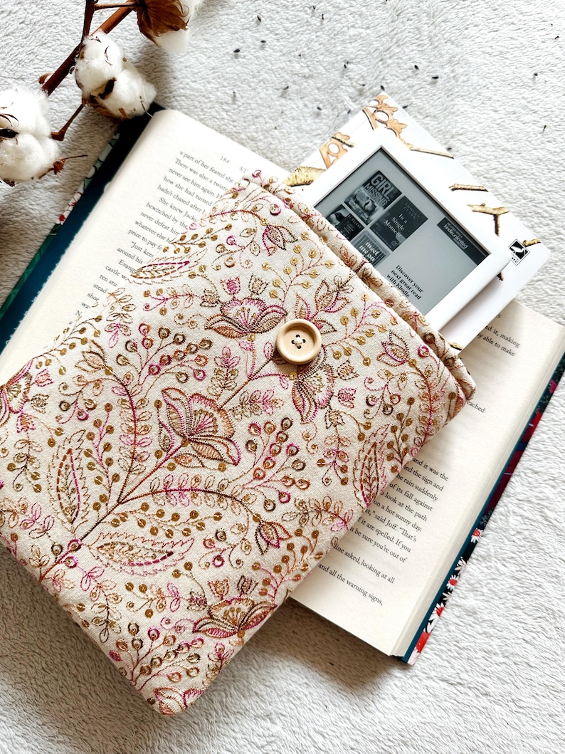 May include: A beige fabric e-reader case with a floral pattern in shades of pink and gold. The case has a button closure and is shown with an e-reader inside.