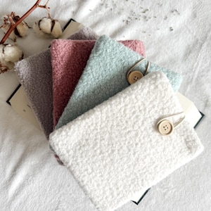 May include: A selection of soft, textured fabric pouches in pastel shades: white, light blue, pink, and grey. Each pouch features a wooden button and loop closure. The pouches are displayed on a white, textured surface.