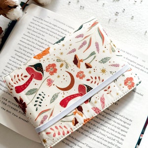 May include: A white book cover with a colourful floral pattern featuring red mushrooms, green leaves, and pink flowers. The cover has a white elastic band and the text "SE FOR TRUE LOVE" is visible on the right side of the cover.