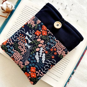 以下が含まれることがあります： 花柄の刺繍が施された濃い青色の布製のポーチ。ポーチには黒い紐の留め具と木製のボタンが付いています。ポーチは本のの上に置かれています。