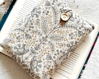 May include: A white book cover with a floral pattern in gold and gray embroidery. The cover has a button closure and a loop for a bookmark.