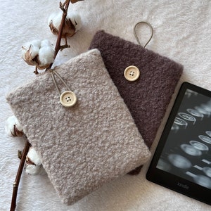 Puede incluir: Dos fundas para Kindle de tela afelpada, una beige y otra marrón, con cierre de botón y un lazo para una correa. Las fundas están sobre una superficie blanca afelpada con una rama de algodón.