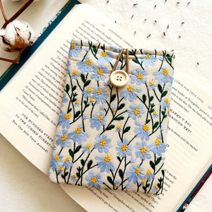 Puede incluir: Una bolsa de tela beige con un dise&ntilde;o de bordado floral azul. La bolsa tiene una correa de cuero marr&oacute;n y un cierre de bot&oacute;n de madera. La bolsa est&aacute; sobre un libro abierto con texto sobre un bosque maldito.