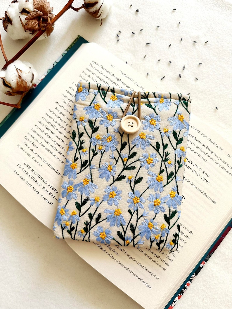 Puede incluir: Una bolsa de tela beige con un dise&ntilde;o de bordado floral azul. La bolsa tiene una correa de cuero marr&oacute;n y un cierre de bot&oacute;n de madera. La bolsa est&aacute; sobre un libro abierto con texto sobre un bosque maldito.