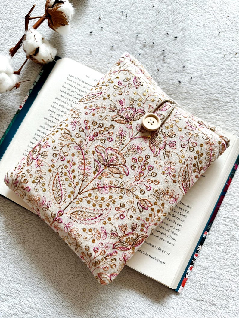 May include: A beige book cover with a floral pattern in pink and gold. The cover has a button closure and a fabric bookmark.