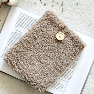 May include: A light brown fluffy book cover with a wooden button and a brown leather strap. The cover is lying on top of a book.