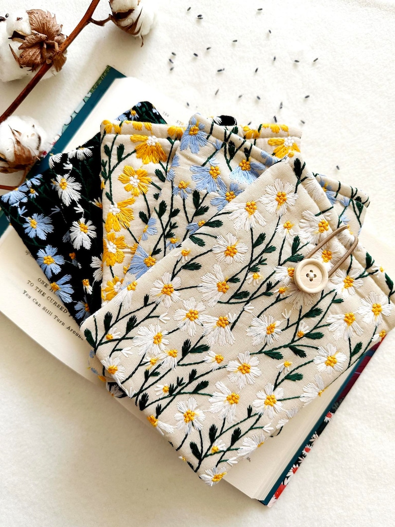 Puede incluir: Cuatro bolsas de tela diferentes con patrones de margaritas bordadas. Las bolsas est&aacute;n apiladas una encima de la otra, con la bolsa superior de color crema con margaritas blancas y tallos verdes. La bolsa de abajo es azul claro con margaritas blancas y tallos verdes. La siguiente bolsa es amarilla con margaritas blancas y tallos verdes. La bolsa inferior es negra con margaritas blancas y tallos verdes.