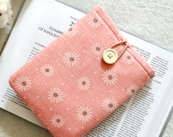 Puede incluir: Una funda de libro de tela rosa con un estampado de margaritas y cierre de botón. La funda está sobre un libro abierto. La funda del libro mide aproximadamente 20 cm por 15 cm.