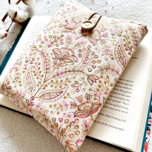 May include: A white book cover with a floral pattern in pink and gold. The cover has a button closure and is made of a soft fabric.