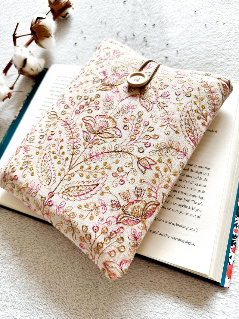 May include: A white book cover with a floral pattern in pink and gold. The cover has a button closure and is made of a soft fabric.