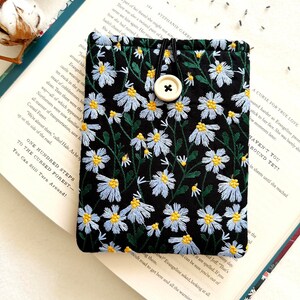 Puede incluir: Una bolsa de tela negra con cierre de bot&oacute;n blanco y un dise&ntilde;o de bordado floral. La bolsa est&aacute; sobre un libro con el texto "One Hundred Steps to the Cursed Forest - You Can Still Turn Around!" impreso en la p&aacute;gina.