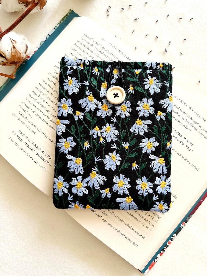 Puede incluir: Una bolsa de tela negra con cierre de bot&oacute;n blanco y un dise&ntilde;o de bordado floral. La bolsa est&aacute; sobre un libro con el texto "One Hundred Steps to the Cursed Forest - You Can Still Turn Around!" impreso en la p&aacute;gina.