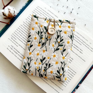 Puede incluir: Una bolsa de tela beige con un dise&ntilde;o bordado de margaritas blancas. La bolsa tiene un cierre de bot&oacute;n marr&oacute;n y un lazo de cord&oacute;n marr&oacute;n. La bolsa est&aacute; sobre un libro abierto con texto sobre un bosque maldito.