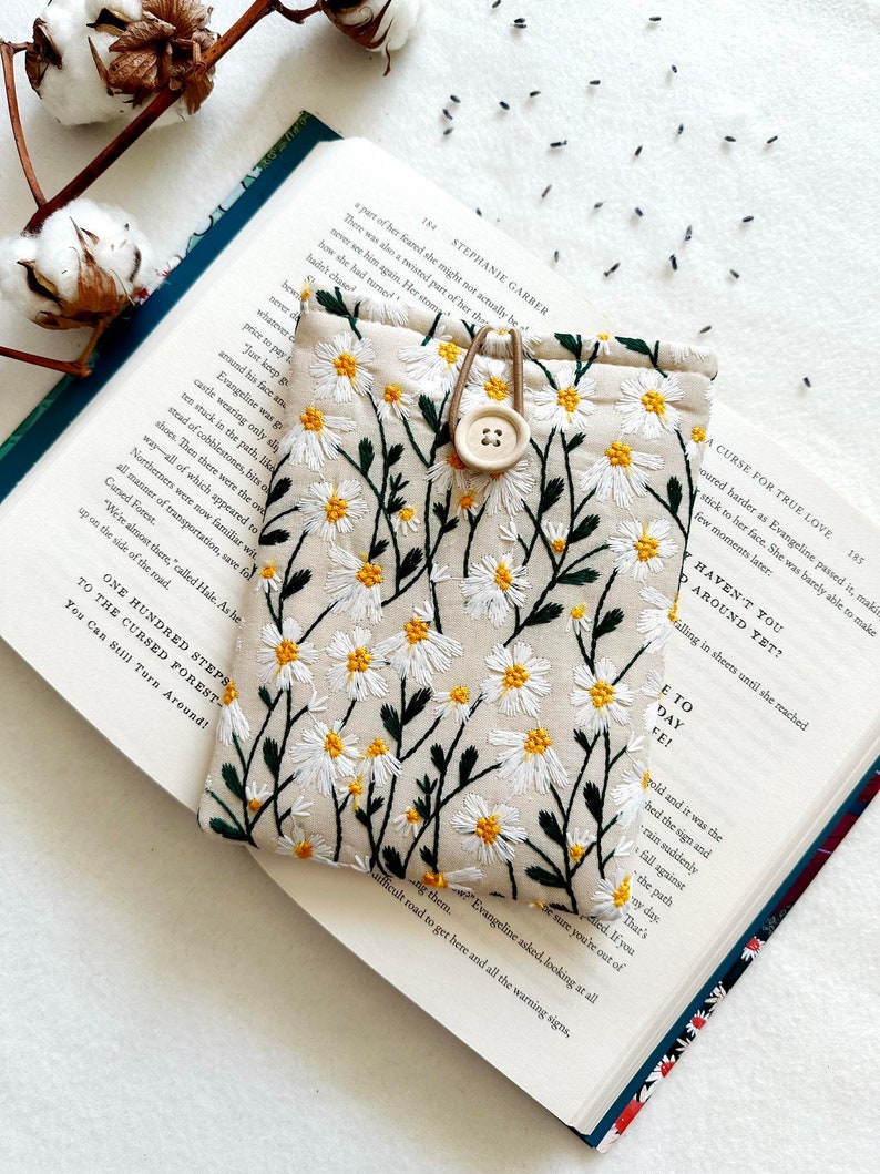 Puede incluir: Una bolsa de tela beige con un dise&ntilde;o bordado de margaritas blancas. La bolsa tiene un cierre de bot&oacute;n marr&oacute;n y un lazo de cord&oacute;n marr&oacute;n. La bolsa est&aacute; sobre un libro abierto con texto sobre un bosque maldito.