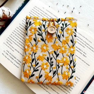 Puede incluir: Una peque&ntilde;a bolsa de tela beige con un dise&ntilde;o de bordado floral amarillo. La bolsa tiene un cierre de bot&oacute;n marr&oacute;n y est&aacute; sobre un libro abierto con texto sobre un bosque maldito.
