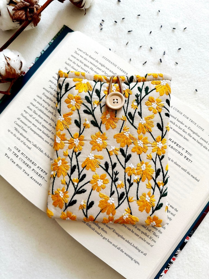 Puede incluir: Una peque&ntilde;a bolsa de tela beige con un dise&ntilde;o de bordado floral amarillo. La bolsa tiene un cierre de bot&oacute;n marr&oacute;n y est&aacute; sobre un libro abierto con texto sobre un bosque maldito.