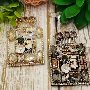 May include: Two decorative perfume bottle-shaped brooches. One is white and gold, the other is black and gold. Both are embellished with various gemstones, beads, and a butterfly design. The brooches are set against a wooden background.