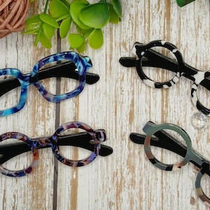 May include: Four pairs of eyeglasses with unique patterned frames. The frames feature various colors, including blue, purple, black, white, and brown. The glasses have black arms and are arranged on a light wood-grain surface.