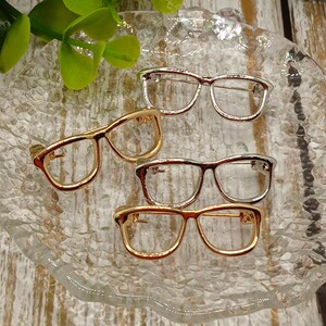 May include: Four pairs of eyeglasses-shaped brooches in gold, silver, and copper tones are arranged on a clear, textured glass plate. The brooches have a classic rectangular frame design, showcasing a variety of metallic finishes. The background is a light-colored wooden surface.