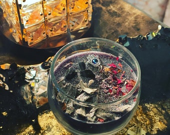 May include: A clear glass candle jar with a purple candle inside. The candle has glitter and dried flowers and herbs inside. A silver charm with a blue eye is on top of the candle.