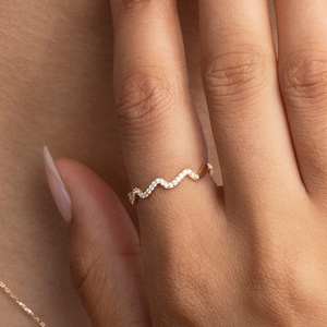 May include: A gold-colored ring with a wavy design, adorned with small, sparkling stones. The ring is on a person's finger, with a gold necklace visible in the background. The ring's design is a continuous wave pattern.