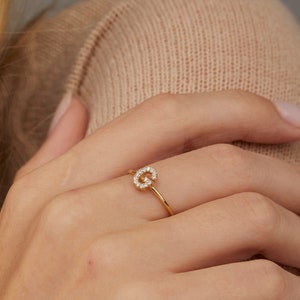 Może przedstawiać: Złoty pierścionek z pojedynczą literą inicjału ozdobioną małymi diamentami. Pierścionek jest noszony na palcu.