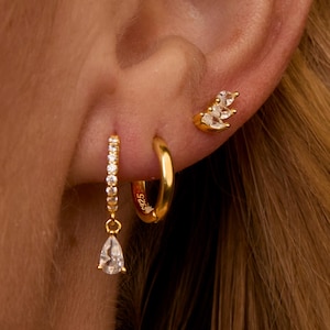 May include: A close-up of an ear adorned with three gold earrings. One is a small stud with a cluster of clear stones. Another is a small gold hoop. The third is a gold hoop with a row of clear stones and a teardrop-shaped clear stone pendant.