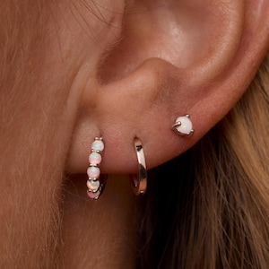 May include: An ear with three earrings: a small white gemstone stud, a silver hoop, and a hoop with white gemstones. The earrings are set against a skin tone background. The jewellery is simple and elegant, suitable for everyday wear.