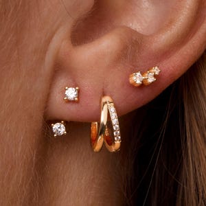 Könnte beinhalten: Nahaufnahme eines Ohrs, das mit mehreren goldenen Ohrringen geschmückt ist. Die Ohrringe umfassen kleine quadratische Ohrstecker, Creolen mit eingebetteten Edelsteinen und eine Ansammlung von drei kleinen, runden Ohrringen.