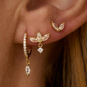 Könnte beinhalten: Goldfarbene Ohrringe mit klaren, funkelnden Akzenten. Das Set beinhaltet einen kleinen Creolen-Ohrring mit einem tropfenförmigen Anhänger, einen blütenförmigen Ohrstecker und einen kleinen Ohrstecker. Die Ohrringe werden an einem Ohr getragen.