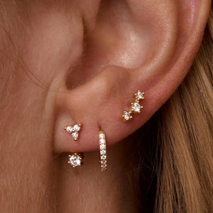 May include: A close-up of an ear adorned with multiple gold earrings. The earrings include small studs with clear gemstones, a small hoop earring, and a cluster of three gemstones. The earrings are set against the backdrop of a person's skin and hair.