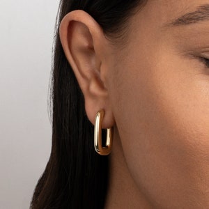 May include: Gold hoop earrings with a rectangular shape. The earrings are worn by a person with dark hair.