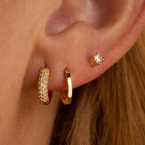 May include: Close-up of a person's ear adorned with three gold earrings. The set includes a small square stud with a clear stone, a small plain hoop, and a larger hoop encrusted with small, sparkling stones. The earrings are set against the backdrop of the person's skin and hair.