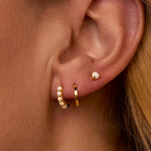 May include: Close-up of a person's ear adorned with gold earrings. The ear features a small stud earring with a white stone, a small gold hoop, and a larger hoop earring with white stones. The earrings are set against the backdrop of the person's skin and hair.