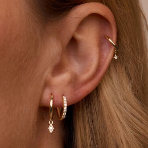 Könnte beinhalten: Nahaufnahme eines Ohrs, das mit mehreren goldenen Ohrringen geschmückt ist. Die Ohrringe umfassen kleine Creolen, einen Reifen mit einem rautenförmigen Design und einen Reifen mit einem hängenden rautenförmigen Anhänger. Ein sternförmiger Ohrring ist ebenfalls sichtbar.