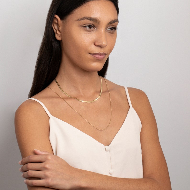 May include: A woman wearing a white tank top with two gold necklaces. The top necklace is a thicker, flat chain, and the bottom necklace is a thinner, round chain.