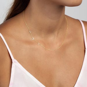 May include: A gold-toned necklace featuring a delicate chain with three small, uppercase letter charms spelling out the name LVY positioned asymmetrically along the right side of the collarbone area, resting against the neck.