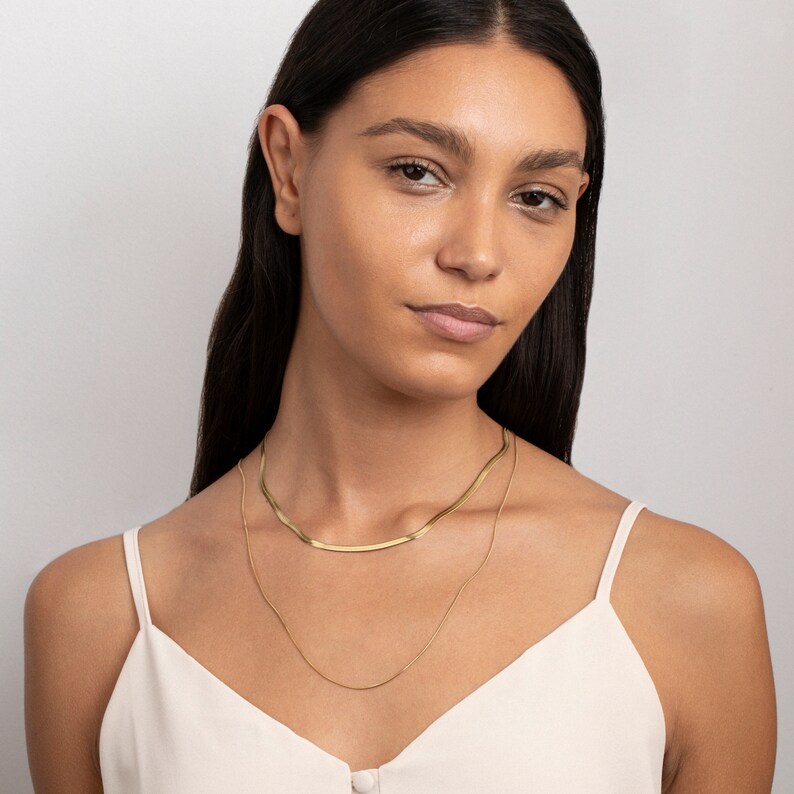 May include: A woman wearing a white tank top with a gold chain necklace layered over a thicker gold chain necklace.